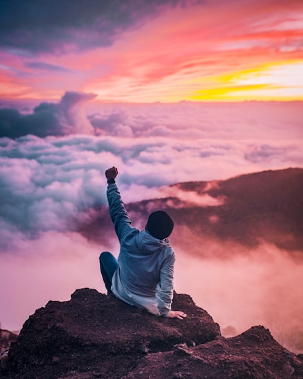 A person with one arm raised sitting on top of a mountain before a gorgeous pink sunset.