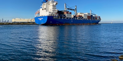 A big container ship stacked with boxes next to the wharf with big cranes and containers stacked on land.