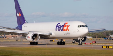 A white FedEx truck on the tarmac behind a large military cargo jet on a cloudy day.